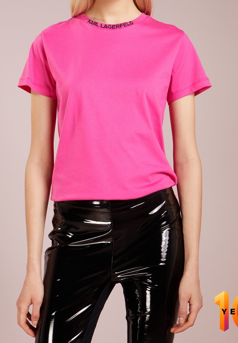 Rosa Baumwoll-T-Shirt mit kurzen Ärmeln und rundem Ausschnitt, mit einem schwarzen "KARL LAGERFELD" Logo am Kragen. Glänzende schwarze eng anliegende Hose.