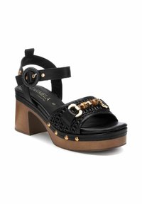 Sandalias de plataforma de cuero negro con una correa trenzada, acentos de hardware dorado y un tacón de madera grueso. Presenta detalles con tachuelas en las correas.