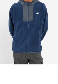 Marinblå fleecetröja med en grå panel på bröstet, hel dragkedja framtill och en huva. Har New Balance-logotypen i vitt.