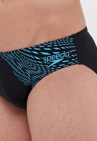 Tanga de natação preta com um padrão gráfico azul com linhas diagonais. O logo Speedo é visível na cintura em azul claro.