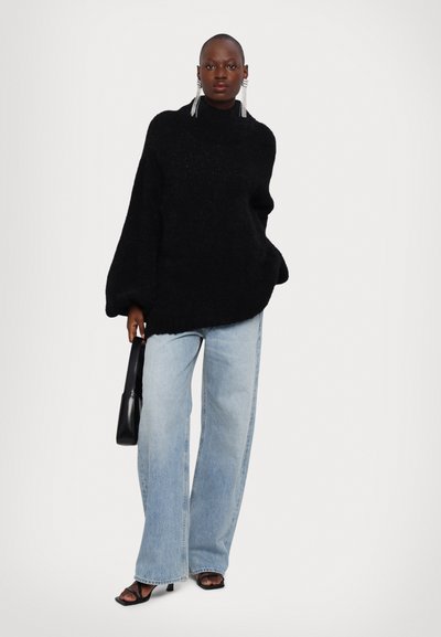 Črna oversized pletena turtleneck pulover s teksturirano površino, v kombinaciji s svetlobluzimi širokimi hlačami in črnimi sandali s peto.