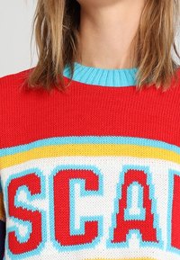 Kobieta ubrana w czerwony dzianinowy sweter z niebieskim kołnierzem i dużymi kolorowymi literami „SCA” widocznymi z przodu.