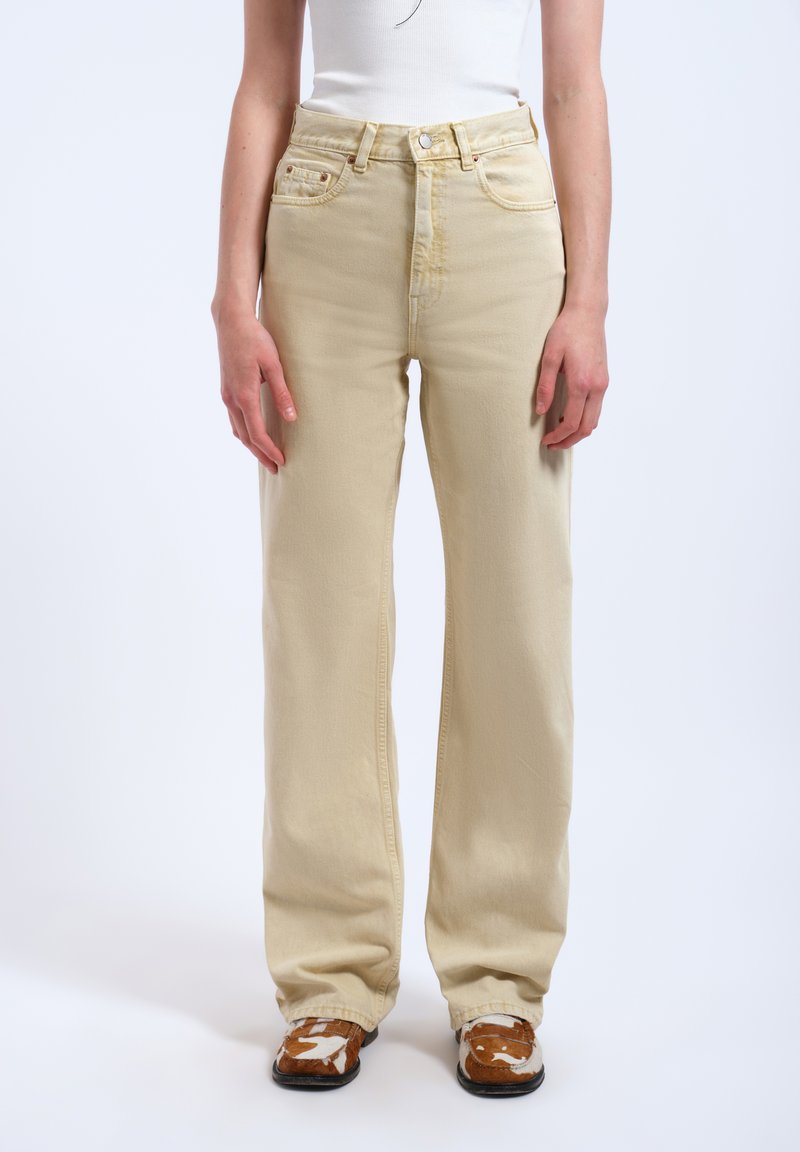 Persoon draagt beige jeans met wijde pijpen, wit mouwloos topje en bruin-witte schoenen met patroon, staande tegen een effen achtergrond.