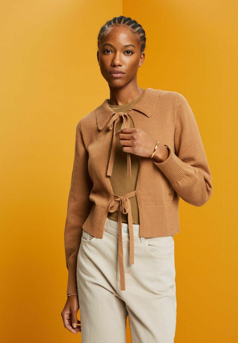 Cardigan court marron avec un col et une fermeture en nœud, associé à un haut beige à côtes et un pantalon beige clair sur un fond jaune vif.