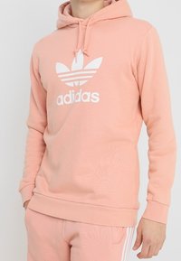 Ljusrosa hoodie i mjukt tyg, med en vit Adidas-logotyp på bröstet, snörning i huvan och ribbade ärmslut och nederkant.
