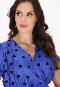 Blaues Kleid aus strukturiertem Satin mit schwarzen, herzförmigen Mustern, V-Ausschnitt und kurzen Ärmeln. Goldene Creolen werden getragen.
