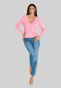 Rosa Cardigan mit V-Ausschnitt, langen Ärmeln und Knopfverschluss, kombiniert mit blauen Jeans und gemusterten High Heels. Einfaches Design, weicher Stoff.