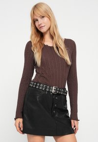 Καφέ μπλούζα με υφή και μακριά μανίκια, συνδυασμένη με μαύρη μίνι φούστα από ψεύτικο δέρμα. Η φούστα έχει κουμπιά μπροστά και ζώνη με τρουκς και χυλοπίτες.