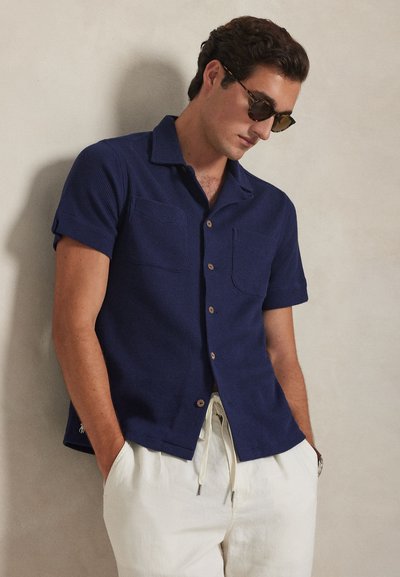 Άνδρας με γυαλιά ηλίου σε σχέδιο χελώνας, μπλε ναυτικό πουκάμισο με κοντά μανίκια και λευκό παντελόνι με κορδόνι, στέκεται μπροστά σε ανοιχτό μπεζ τοίχο.