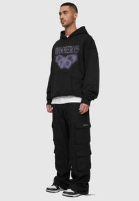 Felpa nera oversize con graphic di farfalla viola, abbinata a pantaloni cargo neri con più tasche, e sneakers nero e bianche.