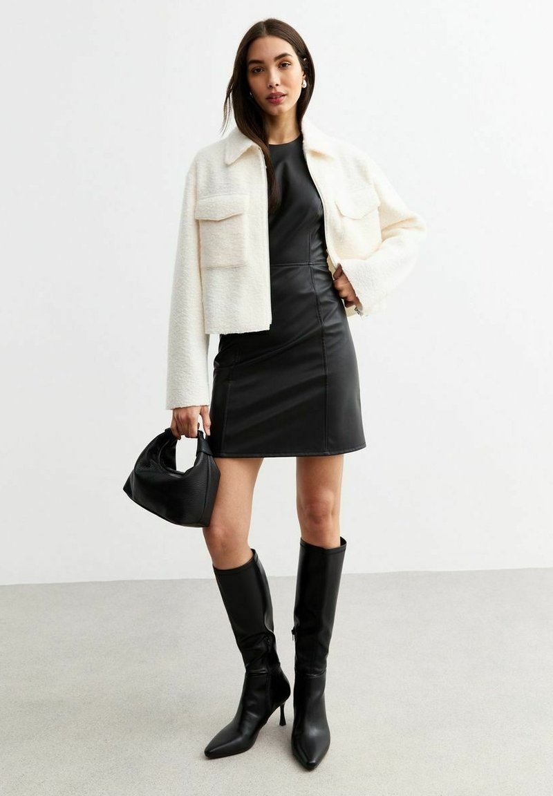 Donna che indossa un giacca bianca a trama sopra un vestito mini in pelle nera, con una borsa nera in mano e stivali alti fino al ginocchio neri con tacco.