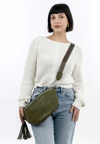 Green leather crossbody bag with a textured finish, zip closure, and a tassel accent. Worn with a white knit sweater and high-waisted jeans.