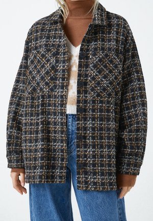 Frau trägt eine schwarz-weiß-braun karierte Jacke mit Knöpfen über einem braunen Rollkragenpullover und einem hell gemusterten Pullover sowie blauen Jeans.