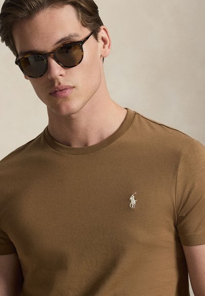 Καφέ βαμβακερό μπλουζάκι με στρογγυλή λαιμόκοψη και κοντά μανίκια, με μικρό λευκό κεντημένο λογότυπο στο στήθος.