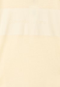 Beige t-skjorte med en glatt tekstur og et sentrert navn i en litt mørkere nyanse. Enkelt, minimalistisk design med korte ermer.