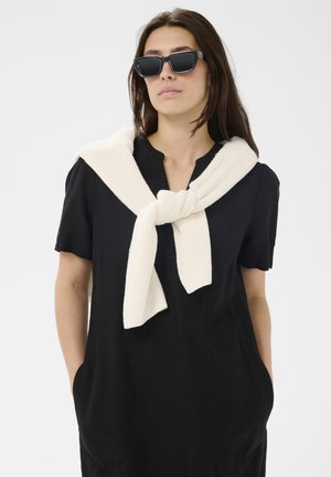 Frau trägt schwarzes Kleid mit Taschen, cremefarbenen Strickpullover über den Schultern gebunden und große schwarze Sonnenbrille, steht vor weißem Hintergrund.