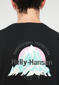 T-shirt preta com um gráfico de montanha em turquesa e branco, rodeado por um círculo rosa e o texto: "Helly-Hansen Proteção Profissional Desde 1877."