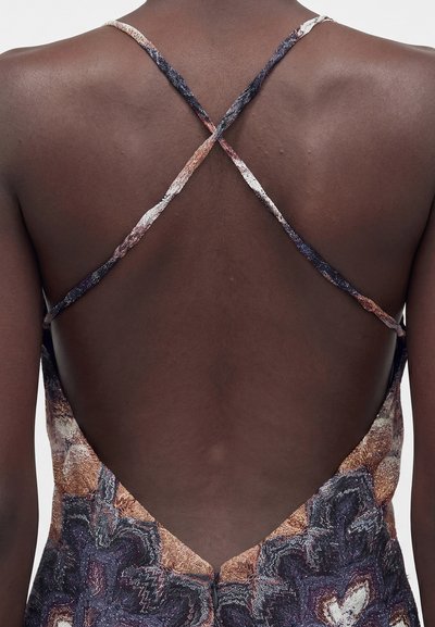 Πλάγια άποψη ατόμου που φοράει φόρεμα με σχέδιο, λεπτές, σταυρωτές τιράντες και ανοιχτή κάτω πλάτη.