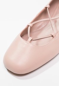 Vaaleanpunaiset nahka flatsit, joissa on neliskulmainen varvasosa. Ominaisuuksiin kuuluvat ristikkäiset hihnat ja sileä, mattapintainen pinta. Vähäistä ompelutyötä näkyvissä.