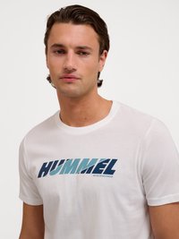 Giovane uomo con capelli corti e scuri che indossa una maglietta bianca con il logo "HUMMEL" e la scritta "Active Sportswear" sul davanti.