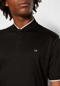Polo noir avec un col mandarin, orné d'un liseré blanc et d'un petit logo "CK" blanc brodé sur le côté gauche de la poitrine. Texture de tissu lisse.