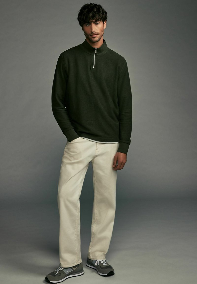 Dunkelgrüner Langarmpullover mit einem Reißverschlusskragen, kombiniert mit hellbeigen Hosen und grauen Sportschuhen. Einfaches und funktionales Design.