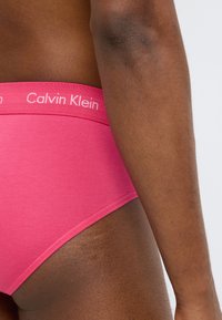 Ροζ βαμβακερό σλιπ Calvin Klein με ομαλή υφή και φαρδιά λευκή ζώνη μέσης με λογότυπο, απεικονιζόμενο από την πλευρά σε ουδέτερο φόντο.
