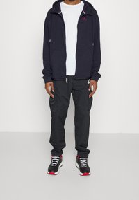 Granatowa bluza z kapturem na zamek z logo, biała koszulka, ciemne spodnie cargo i czarno-czerwone buty sportowe. Posiada kieszenie oraz fakturowany materiał.