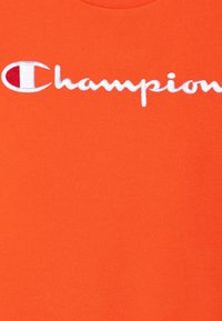 T-shirt di cotone arancione con un grande logo "Champion" ricamato in bianco, con un distintivo accento rotondo rosso sulla lettera "C".