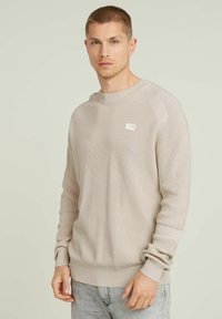 Maglione beige a coste con scollo rotondo e maniche raglan. Presenta una piccola etichetta con il marchio sul petto. Il tessuto ha una finitura texturizzata.