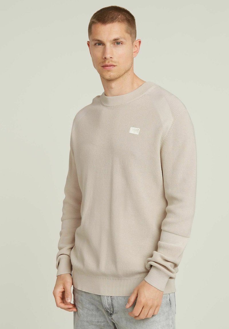 Maglione beige a coste con scollo rotondo e maniche raglan. Presenta una piccola etichetta con il marchio sul petto. Il tessuto ha una finitura texturizzata.