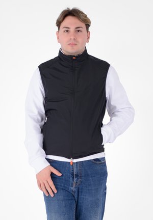 Gilet nero senza maniche con colletto alto, chiusura a zip anteriore e zip arancione, abbinato a una camicia bianca a maniche lunghe e jeans blu.