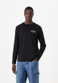 Must mustärk särk pikad varrukad, millel on väike valge "HILFIGER" logo. Kandmine helesiniste cargo teksadega küljetaskutega. Lihtne, igapäevane disain.
