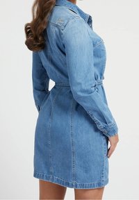 Nainen, jolla on pitkä kihara hius, pukeutunut istuvaan vaaleansiniseen farkkujakkuun, joka näyttää selkä- ja sivuyksityiskohdat nappikiinnityksellä.