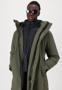 Olivgrüner Parka mit einer schwarzen Innenschicht, ausgestattet mit einer Kapuze, Reißverschlüssen und Knopfverschlüssen. Kombiniert mit dunklen Sonnenbrillen für einen Kontrast.