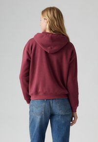 Burgundy pullover hoodie with a drawstring hood, ribbed cuffs, and hem; paired with light blue jeans featuring back pockets.