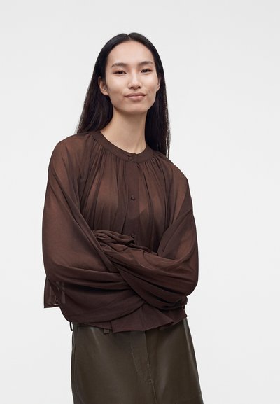 Femme aux longs cheveux foncés portant un chemisier ample, transparent marron foncé et une jupe sombre, debout les bras croisés devant un fond uni.