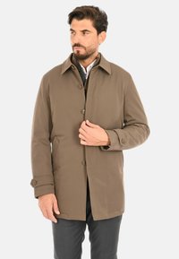 Cappotto marrone con bottoni, collo, maniche lunghe e tasche laterali, realizzato in materiale liscio, indossato sopra una camicia più chiara.