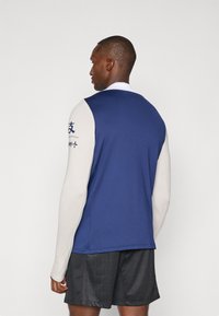 Long-sleeve athletic top with a blue body and cream sleeves, featuring printed text on the left arm and a smooth, stretchy fabric.