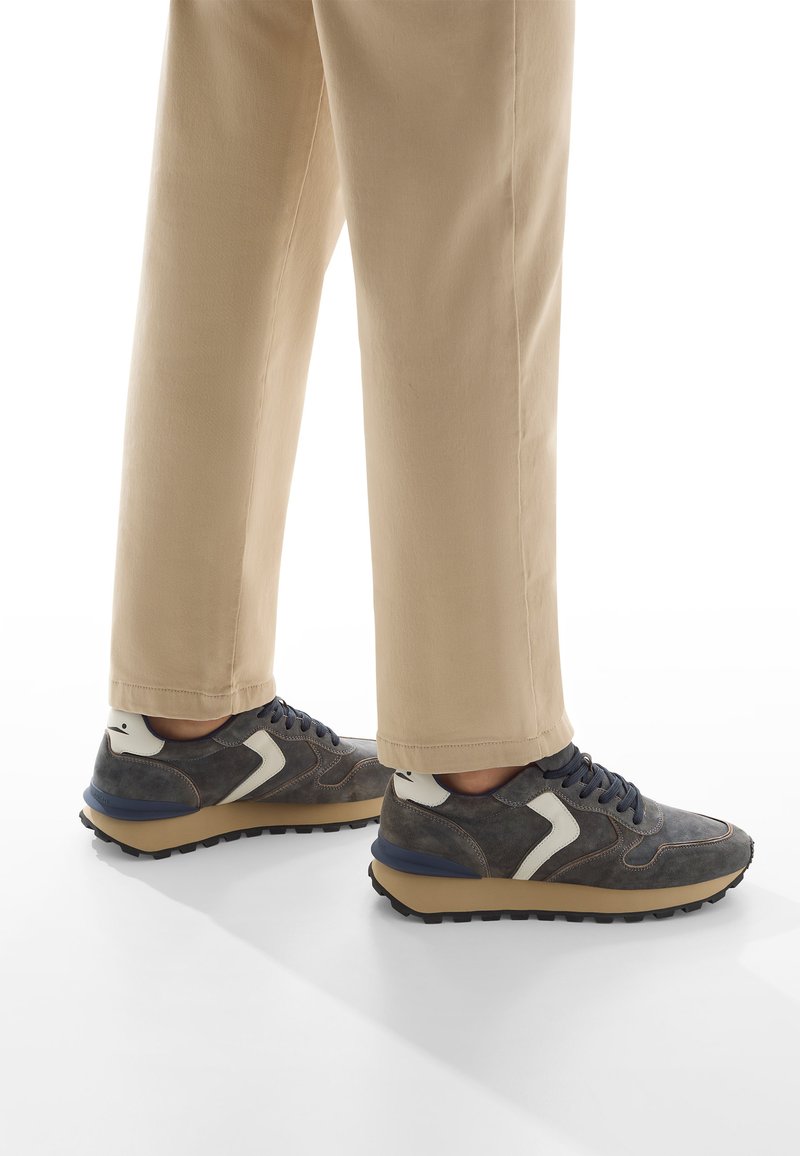 Sneaker grigie con lacci blu navy e dettagli bianchi, caratterizzate da un materiale testurizzato, design a profilo basso e una robusta suola nera.