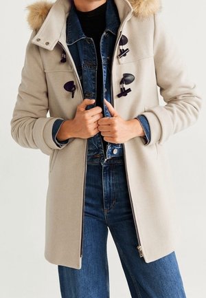 Personne portant un manteau beige avec des boutons à bascule sur une veste en jean bleue et un jean, fermant la fermeture éclair du manteau avec les deux mains.