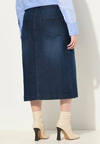 Gonna midi in denim blu scuro con tasche posteriori, taglio dritto e orlo poco sotto il ginocchio, abbinata a stivaletti beige alla caviglia.