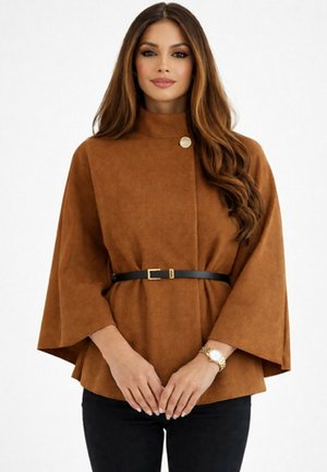 Femme aux longs cheveux bruns portant un manteau style cape marron avec un col haut, une ceinture noire et une montre en or, se tenant devant un fond blanc.