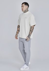 Bež oversized T-särk lühikeste varrukatega, hallid kitsad püksid ja valged tossud. Märgatavad tätoveeringud käte peal ning bränding särgil ja pükstel.