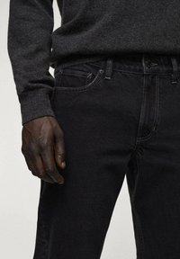 Jeans de mezclilla negra con corte recto, con costuras visibles, un bolsillo frontal y un acabado de textura suave. Acompañados de un suéter gris oscuro.