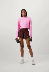 Svetloružový skrátený sweatshirt, tmavohnedé šortky, drží činky. Tenisky s bielymi akcentami; aerodynamický dizajn, hladká textúra.