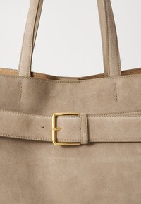Sac cabas en daim beige avec une forme plate et structurée. Doté d'une sangle avec boucle en ton or et de deux poignées robustes. Texture lisse sur toute la surface.