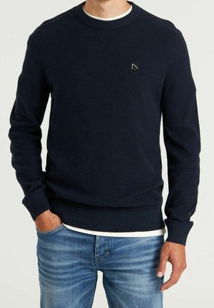 Uomo che indossa un maglione blu navy texturizzato con una piccola toppa logo sul petto, una maglietta bianca e jeans azzurri con la mano in tasca.