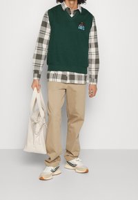 Grön ärmlös tröja över en grå och vit rutigt skjorta, beige byxor och vita sneakers med svarta och orangea accenter. Bär en krämfärgad väska.