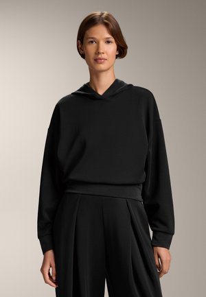 Felpa nera con una vestibilità rilassata, cappuccio con cordino e orlo a coste. Abbinata a pantaloni neri larghi e drappeggiati, creando un look monocromatico.
